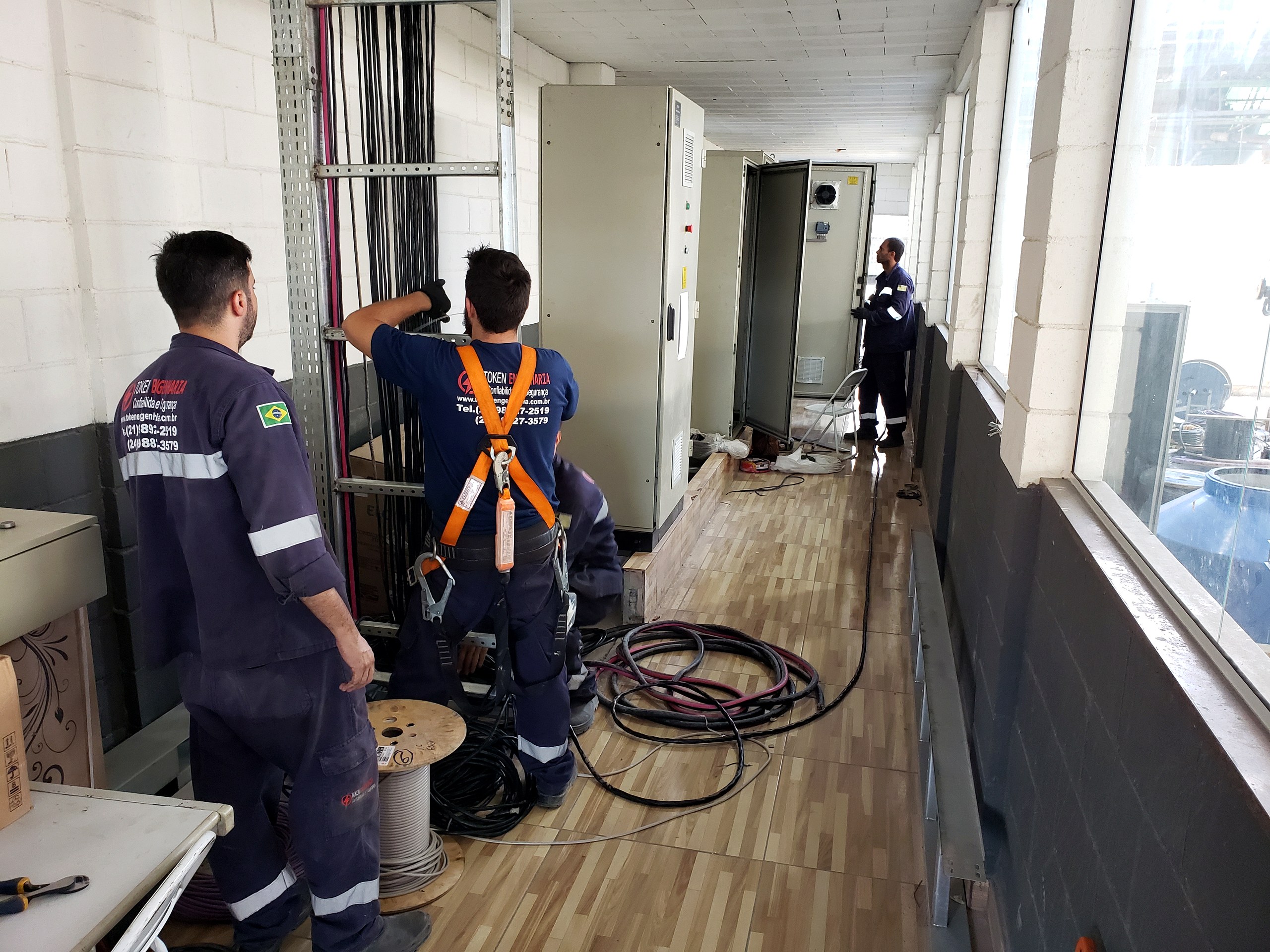 Eletricistas da Token Engenharia lançando cabos em eletrocalha junto a painéis elétricos em área técnica predial