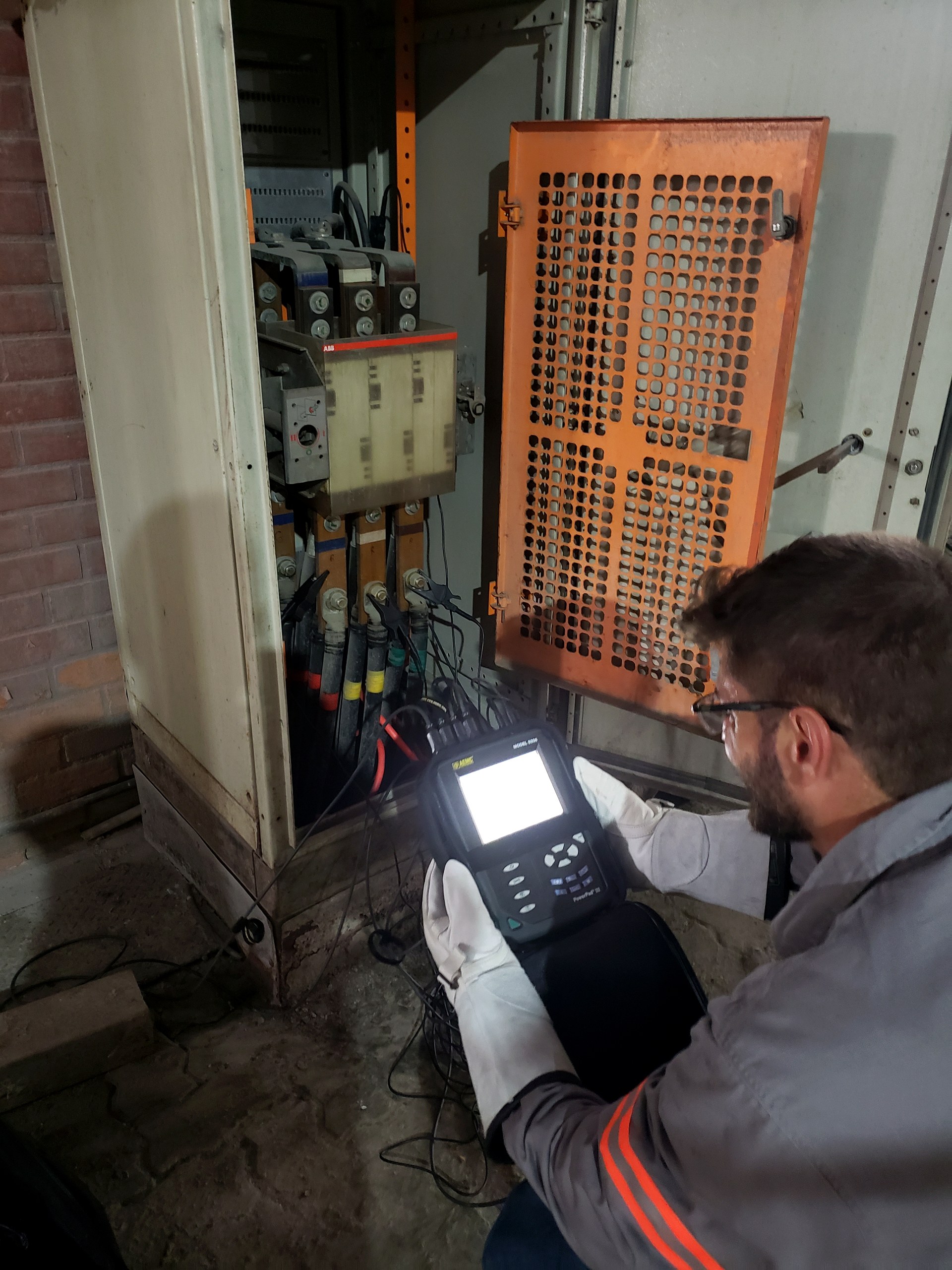Técnico da Token Engenharia com luvas isolantes medindo qualidade de energia em painel elétrico com disjuntor ABB e barramentos de cobre aberto