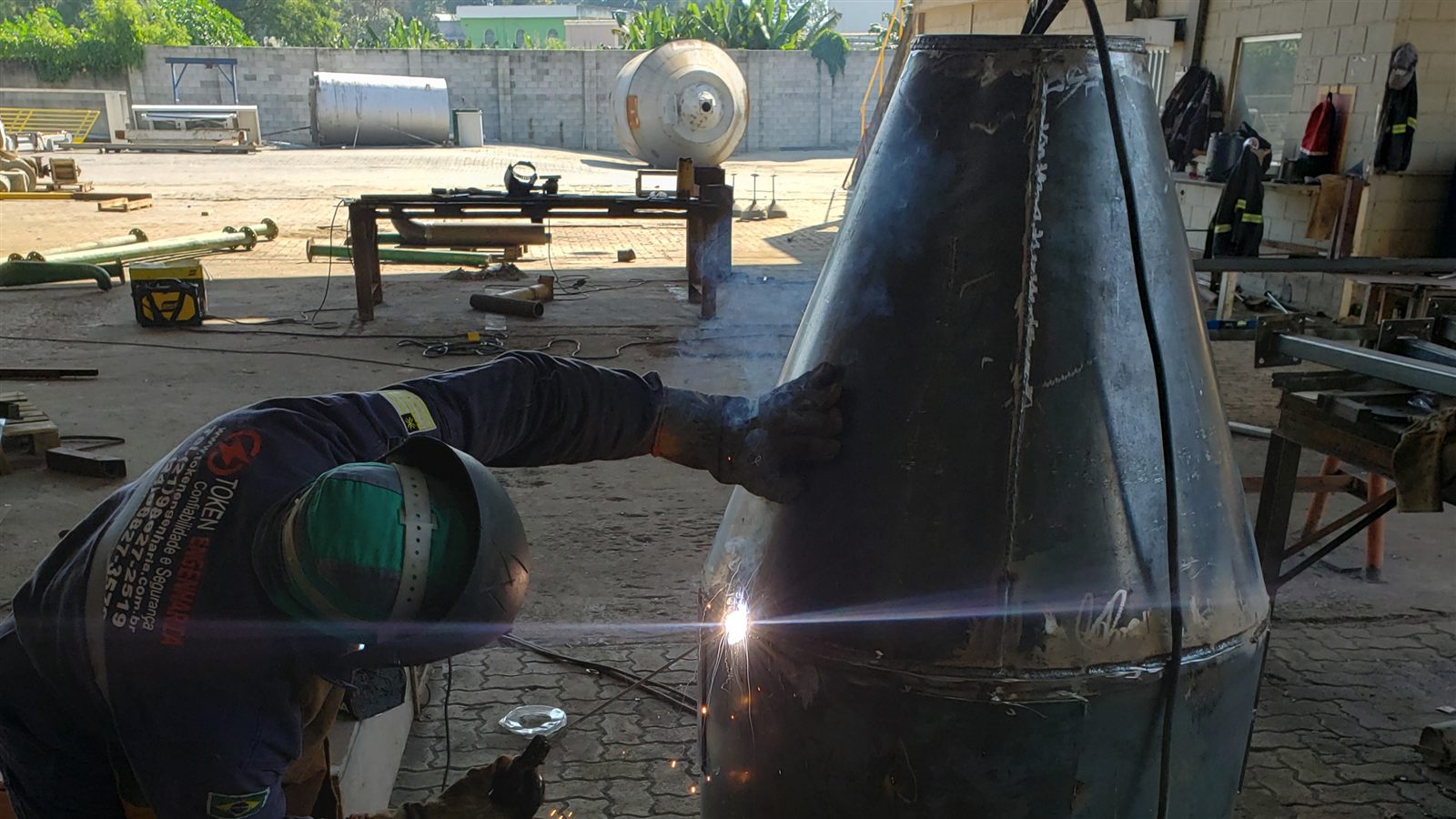 Soldador trabalhando em peça cônica metálica de grande porte em galpão de caldeiraria da Token Engenharia com esmerilhadeira e inversora de solda